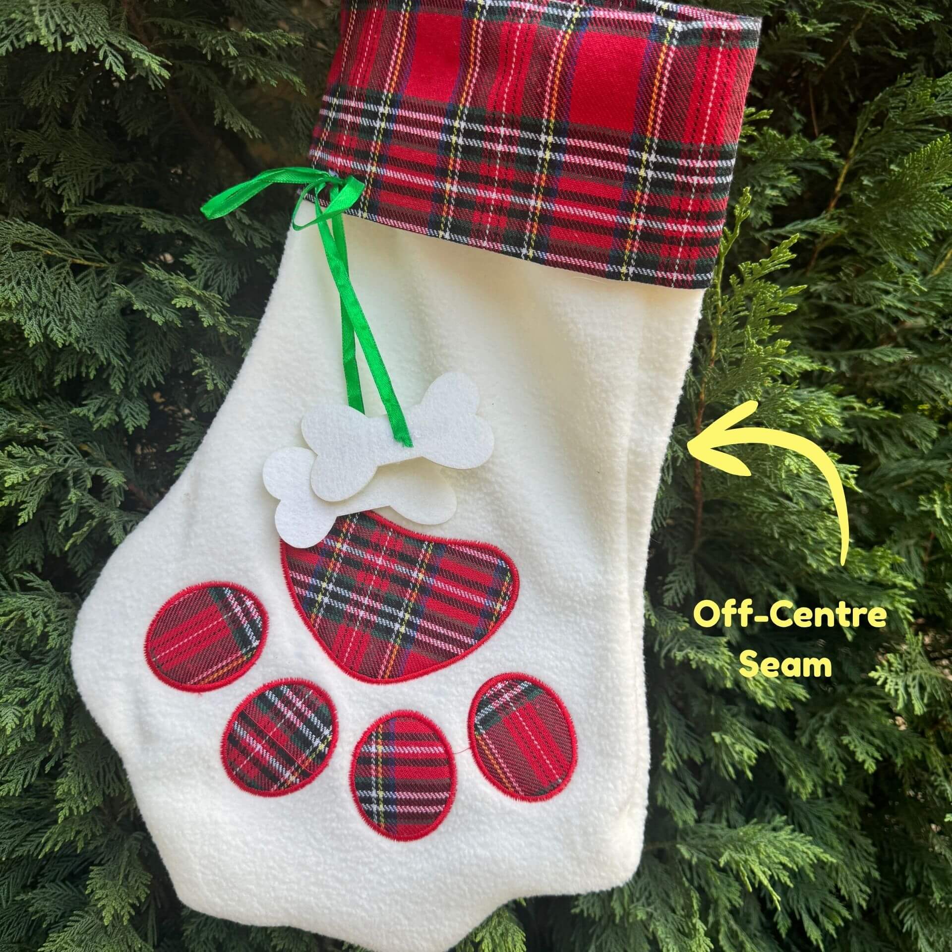 White Christmas stocking with red plaid accents and off-center seam, hanging against a green tree background.